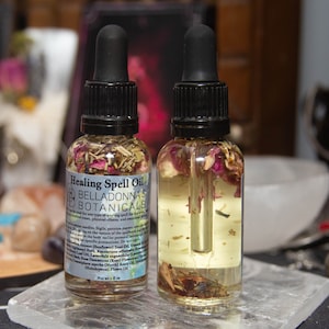 May include: Two small glass bottles filled with a clear liquid and dried herbs and flowers. The bottles have black dropper tops and labels that read "Healing Spell Oil" and "Belladonna's Botanicals".