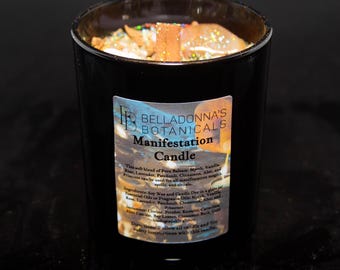 Pode incluir: Um frasco de vela de vidro preto com uma tampa dourada. A vela é etiquetada como "Manifestation Candle" e tem uma descrição dos ingredientes e instruções de uso. A vela é cercada por um fundo abstrato colorido.