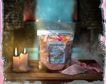 May include: A clear plastic bag filled with pink bath salts and dried rose petals. The bag is labeled "Custom Bath Salts" and "BellaDonna's Botanicals". The bath salts are sitting on a wooden surface with two lit candles.