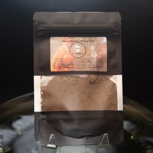 May include: A black pouch containing Santa Muerte Ritual Dirt. The label reads "Santa Muerte Ritual Dirt" with a skull graphic and the brand name "LAMIA". The pouch is held in a clear plastic stand, and the contents are a brown powder.
