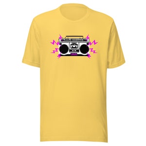 May include: A yellow t-shirt featuring a black and white illustration of a boombox with pink lightning bolts. The boombox has the text "CBZL" on it.