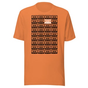 May include: Orange t-shirt featuring a black and white graphic of cassette tapes. One cassette tape is highlighted in orange with the word "CRUZL" printed on it.