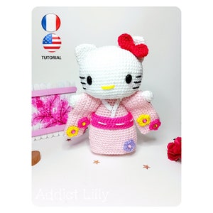 Peut inclure: Une poupée Hello Kitty en crochet portant un kimono rose avec des fleurs jaunes et violettes. La poupée a un visage blanc avec des yeux noirs et un nez jaune. La poupée porte un nœud rouge sur la tête. La poupée est assise sur une surface blanche avec un fond rose et blanc.