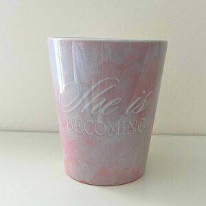 以下が含まれることがあります： ピンクと白の大理石模様のセラミックタンブラー。「She is BECOMING」の文字が前面に白で印刷されています。タンブラーはわずかに先細りの形状をしています。