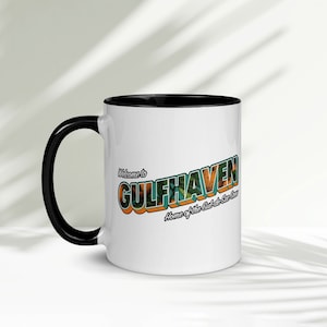 Puede incluir: Taza de café de cerámica blanca con borde y asa negros. La taza presenta un gráfico de estilo retro que dice "Bienvenido a Gulfhaven, Home of the Cut-a-See Sea."