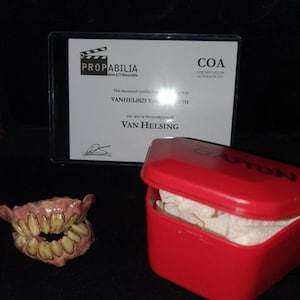 Puede incluir: Un conjunto de dientes falsos en una caja de plástico roja con un certificado de autenticidad. El certificado indica que los dientes se utilizaron en la producción de la película "Van Helsing".