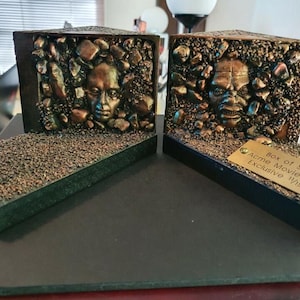 Puede incluir: Dos sujetalibros de bronce con forma de caja con una cara asomando desde cada uno. La caja de la derecha tiene una placa dorada que dice "Box of Souls Acme Movie Store Exclusive 1/15".