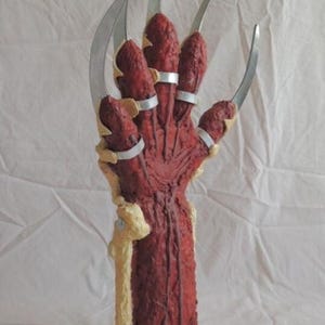 May include: A prop hand with long, curved metal blades extending from the fingers. The hand is a deep red colour with a textured appearance, mounted on a silver base. The text "mr-jvoorhees ebay" is at the bottom.