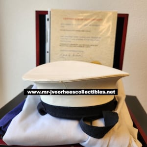 May include: A white naval officer's hat with a black band and brim, resting on a white cloth inside a wooden display case. A document is visible in the background. The website address www.mr-jvoorheescollectibles.net is printed on the image.