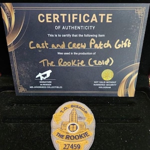 以下が含まれることがあります： 「The Rookie (2010)」の「Cast and Crew Patch Gift」の真正性証明書。パッチは楕円形で、「T.O. Bishop」と「The Rookie 27459」の文字が入っています。証明書は黒色で、金色の文字が書かれています。