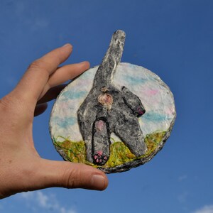 May include: A round, gray, felt sculpture of a cat's rear end with pink paws and a white background with blue and pink clouds.
