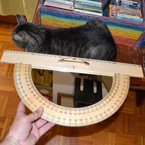 May include: A wooden protractor with a clear plastic protractor attached to it. The protractor is marked with numbers from 0 to 180 degrees. The protractor is being held by a hand.