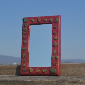 Puede incluir: Espejo rectangular con marco rojo y piedras decorativas verdes y marrones. El espejo se muestra al aire libre sobre un fondo de cielo azul. El marco está hecho de un material texturizado.