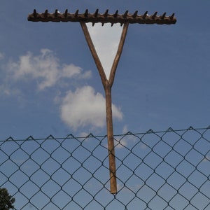 Puede incluir: Un rastrillo de madera con un marco triangular está montado en un poste de madera. El rastrillo está contra un cielo azul con nubes blancas y una valla de cadena verde.