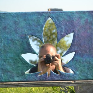 May include: A rectangular artwork with a cannabis leaf cutout, painted in shades of blue and purple. The textured surface reflects the surrounding environment, including a person holding a camera.