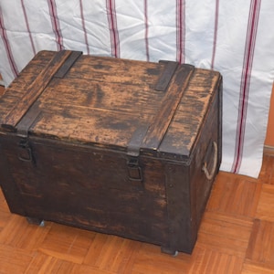 Op de afbeelding: Een donkerbruine houten opbergkist met metalen scharnieren en banden. De kist heeft een versleten, rustieke uitstraling en staat op een houten vloer.