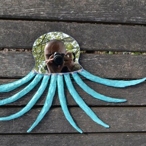 May include: A unique octopus-shaped mirror with a reflective surface and eight turquoise tentacles. The mirror's frame is a light blue color. The mirror is placed on a weathered wooden surface.