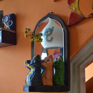 Puede incluir: Un espejo con marco de madera y un estante debajo. El espejo es arqueado en la parte superior y tiene una pantalla de lámpara blanca con un diseño de gato azul en el interior. Tres pequeñas figuritas de gato están en el estante, una negra y amarilla, una naranja y una verde.