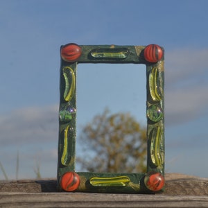 Puede incluir: Un pequeño marco de fotos verde con un espejo en el interior. El marco está decorado con canicas de vidrio rojas y amarillas.