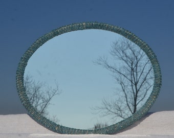 May include: A round, teal-colored mirror with a woven frame sits on a white surface. The mirror reflects a blue sky and a bare tree.