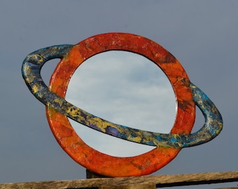 May include: A round mirror with a colorful, planet-like frame. The frame is painted with orange, blue, and gold hues. The mirror is mounted on a wooden structure against a cloudy sky.