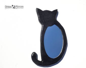 Op de afbeelding: Zwarte kattenvormige wandspiegel met een blauw reflecterend oppervlak. De spiegel is gemaakt van keramiek en heeft een uniek ontwerp.