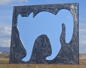 Op de afbeelding: Een zwarte spiegellijst met een uitgesneden katten-silhouet. De spiegel reflecteert de blauwe lucht en witte wolken.