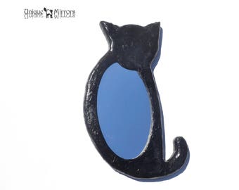 Op de afbeelding: Zwarte kattenvormige spiegel met een blauw reflecterend oppervlak. De spiegel is gemaakt van hout en heeft een gladde, glanzende afwerking.