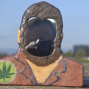 Puede incluir: Un espejo artesanal con forma de cabeza y hombros. El marco es marrón con barba y pelo castaño oscuro. Un círculo verde con una hoja de cannabis está en el hombro. El espejo refleja el entorno.