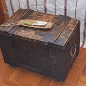 Op de afbeelding: Een donkerbruine houten kist met metalen scharnieren en een touwgreep. De kist staat op een houten vloer.