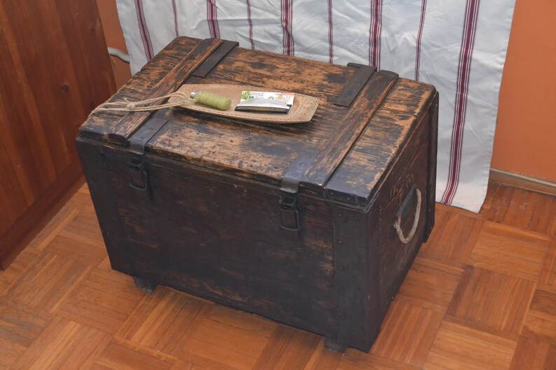 Op de afbeelding: Een donkerbruine houten kist met metalen scharnieren en een touwgreep. De kist staat op een houten vloer.