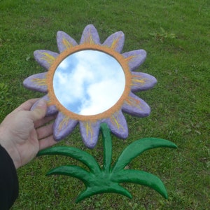 May include: A whimsical, handcrafted flower mirror. The mirror is round, surrounded by a purple flower with yellow accents. The stem and leaves are green. The mirror reflects a cloudy sky.