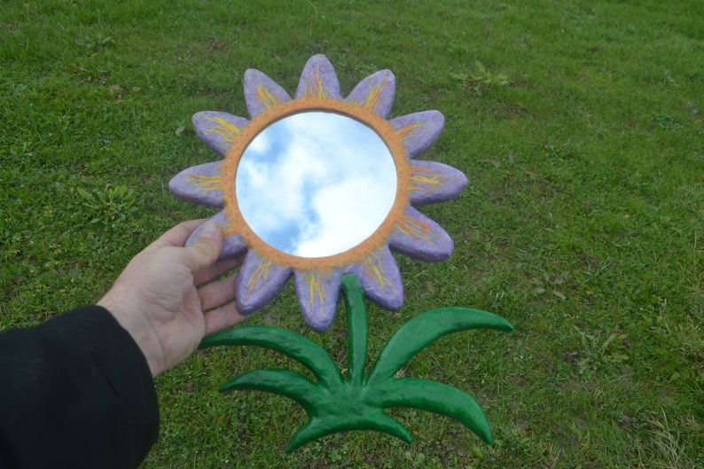 May include: A whimsical, handcrafted flower mirror. The mirror is round, surrounded by a purple flower with yellow accents. The stem and leaves are green. The mirror reflects a cloudy sky.