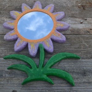 May include: A decorative flower-shaped mirror with a purple petal design, orange center, and yellow accents. The mirror reflects a blue sky with clouds. The flower has a green stem and leaves, all mounted on a wooden surface.
