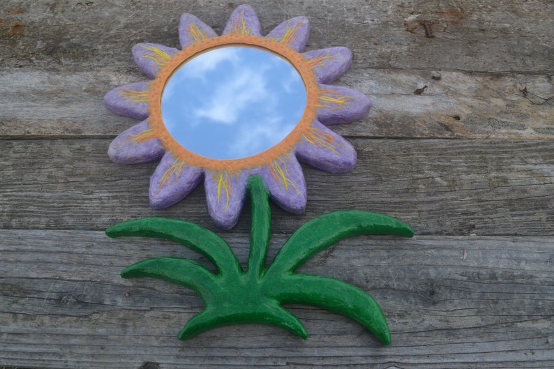May include: A decorative flower-shaped mirror with a purple petal design, orange center, and yellow accents. The mirror reflects a blue sky with clouds. The flower has a green stem and leaves, all mounted on a wooden surface.