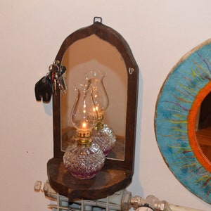 May include: A wooden arched mirror with a shelf underneath. Two oil lamps with clear glass shades and pink bases are on the shelf. The mirror is hanging on a white wall.
