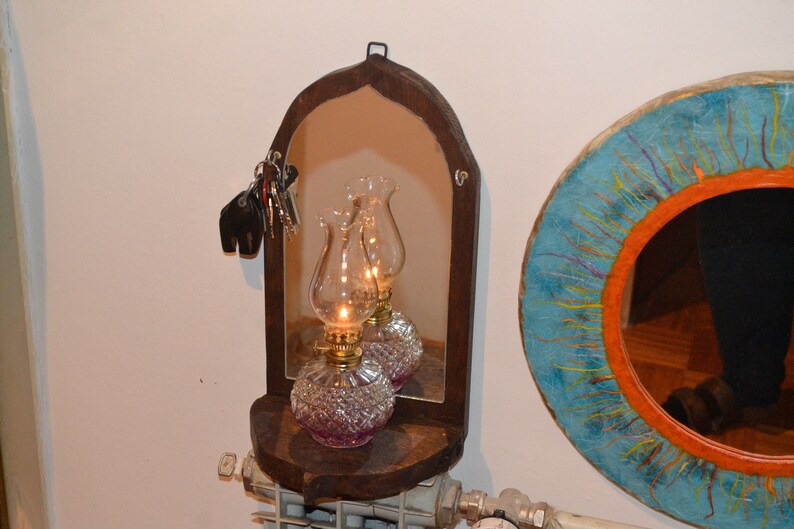 May include: A wooden arched mirror with a shelf underneath. Two oil lamps with clear glass shades and pink bases are on the shelf. The mirror is hanging on a white wall.