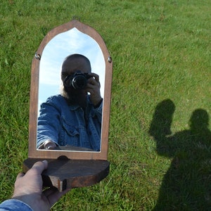 May include: A wooden framed mirror with an arched top and a shelf. A person is reflected in the mirror taking a selfie with a camera.
