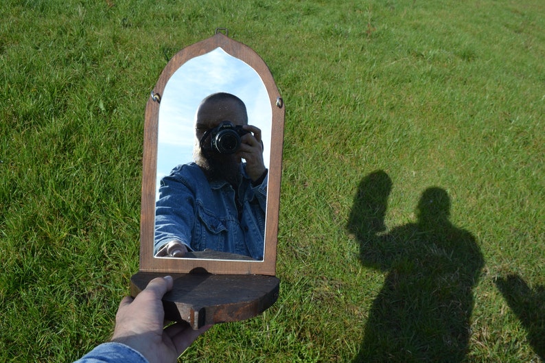 May include: A wooden framed mirror with an arched top and a shelf. A person is reflected in the mirror taking a selfie with a camera.