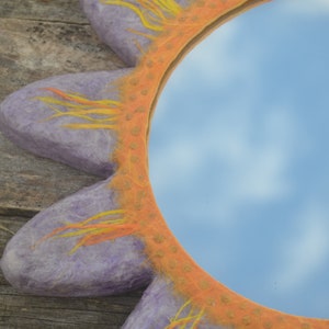 May include: A decorative mirror with a sun-shaped frame. The frame is a light purple color with orange and yellow accents. The mirror reflects a blue sky with clouds.