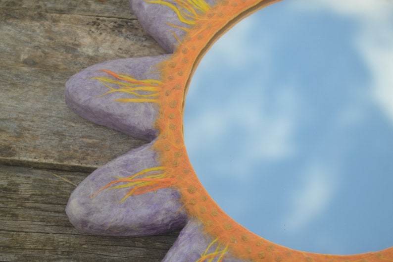 May include: A decorative mirror with a sun-shaped frame. The frame is a light purple color with orange and yellow accents. The mirror reflects a blue sky with clouds.