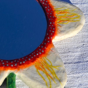 May include: A flower-shaped mirror with a blue reflective surface. The mirror's frame is white with red and orange accents, and small silver dots. Yellow lines extend from the orange areas. A green stem is visible.