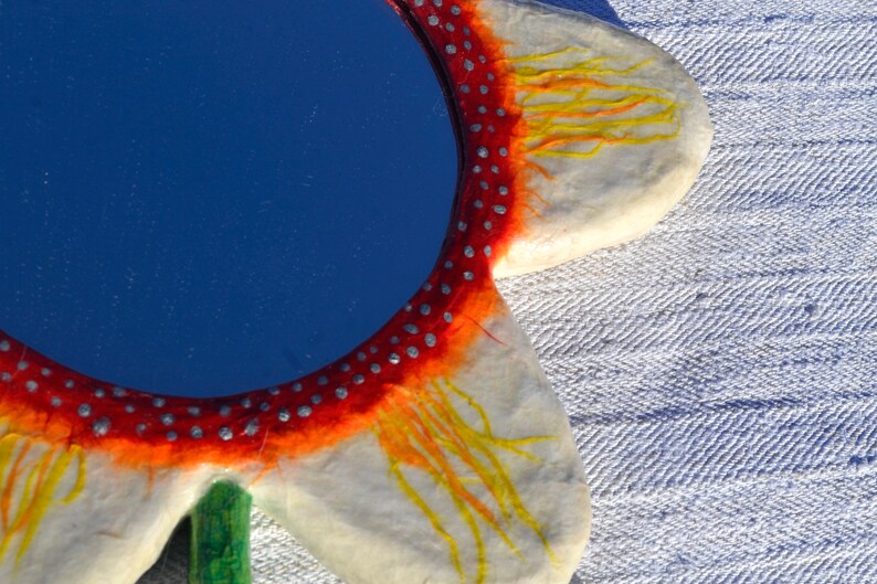 May include: A flower-shaped mirror with a blue reflective surface. The mirror's frame is white with red and orange accents, and small silver dots. Yellow lines extend from the orange areas. A green stem is visible.