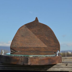 May include: A wooden wall mirror with a hinged top. The mirror is oval shaped and has a dark wood frame. The mirror is closed and the top is angled.