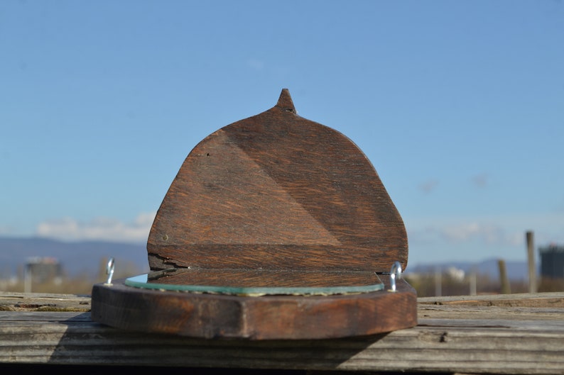 May include: A wooden wall mirror with a hinged top. The mirror is oval shaped and has a dark wood frame. The mirror is closed and the top is angled.