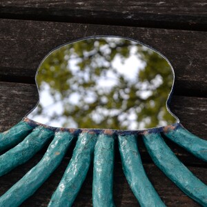 May include: A handmade mirror with a unique octopus design. The mirror has a teal frame with eight textured tentacles extending outwards. The mirror's surface reflects a blurred outdoor scene, and the frame is set against a dark wooden background.