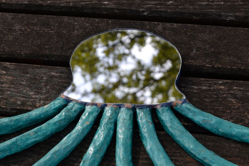 May include: A handmade mirror with a unique octopus design. The mirror has a teal frame with eight textured tentacles extending outwards. The mirror's surface reflects a blurred outdoor scene, and the frame is set against a dark wooden background.