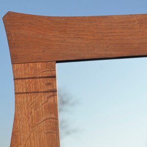 May include: Close-up of the top corner of a wooden mirror frame. The frame is made of light brown wood with a natural grain pattern. The mirror reflects a blue sky and a blurry tree.