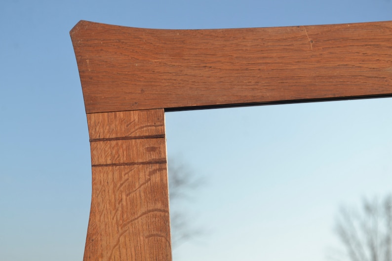 May include: Close-up of the top corner of a wooden mirror frame. The frame is made of light brown wood with a natural grain pattern. The mirror reflects a blue sky and a blurry tree.