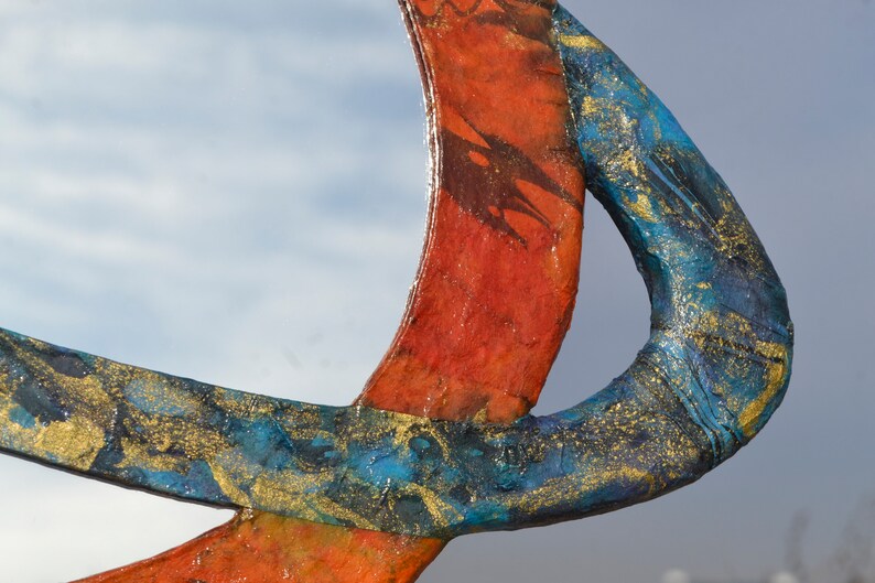 May include: Abstract sculpture with intersecting curved forms. The sculpture features vibrant colors, including red, blue, and gold. The blue section has a textured, speckled appearance with gold accents. The red section has a smooth finish.